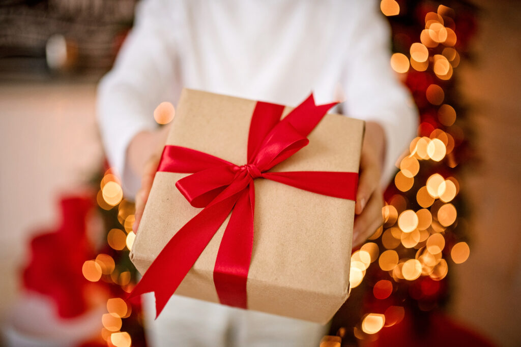 Ręce podające prezent w brązowym papierze przewiązany czerwonym wstążką na tle światełek choinkowych