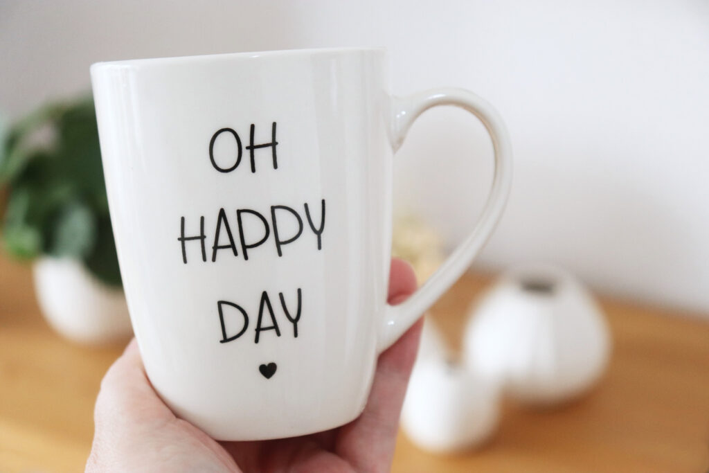 Dłoń trzyma biały kubek z napisem ‘OH HAPPY DAY’ na przytulnym, rozmytym tle domowym