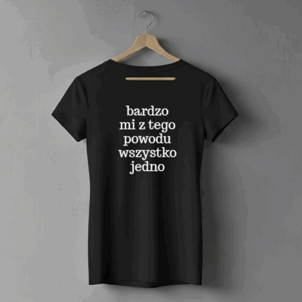 Czarna koszulka na wieszaku wiszącym na szarej ścianie, z białym napisem w kilku linijkach: ‘bardzo mi z tego powodu wszystko jedno’. Koszulka ma prosty krój i krótkie rękawy