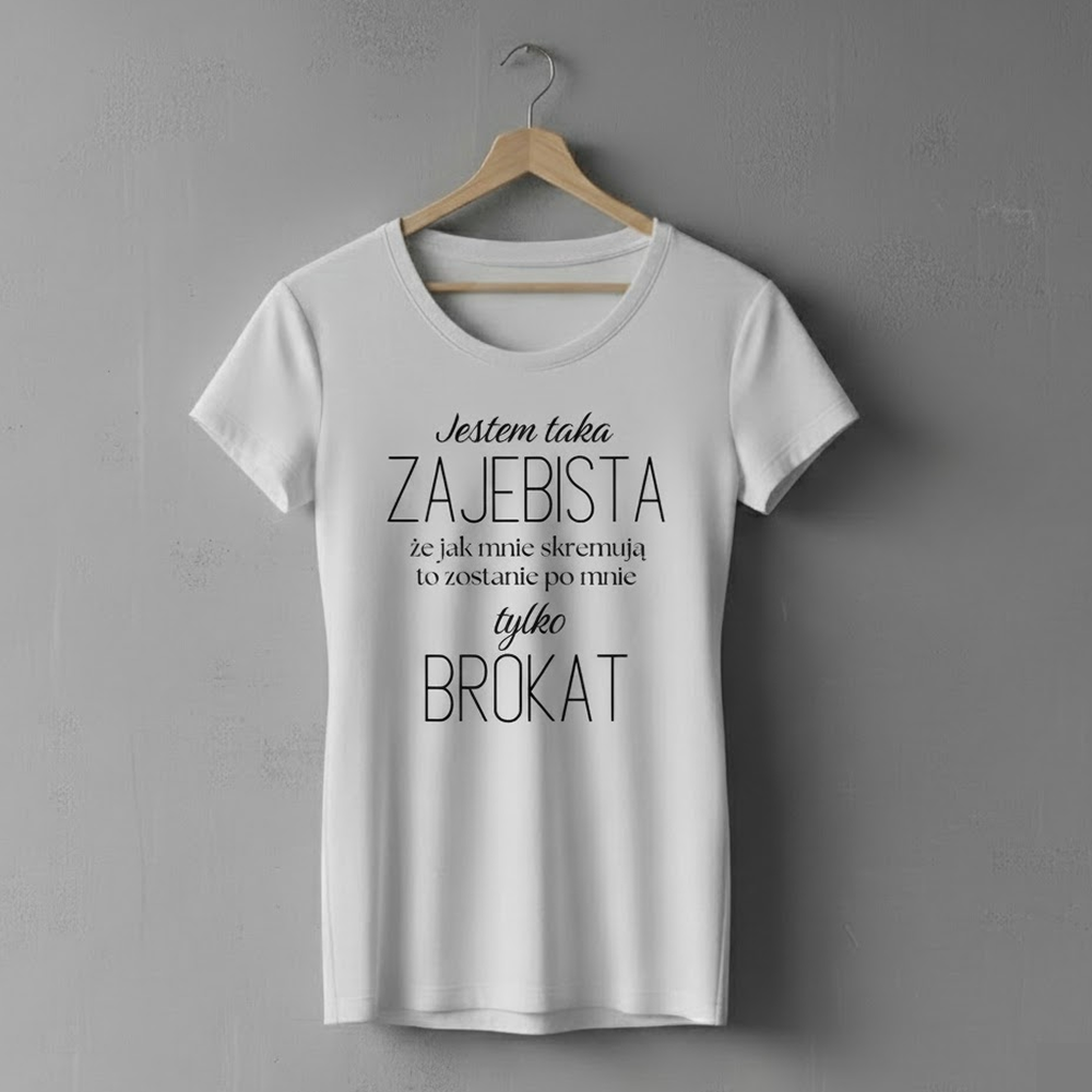41241184 Biała koszulka wisząca na drewnianym wieszaku, z nadrukiem humorystycznego napisu: ‘Jestem taka zajebista, że jak mnie skremują, to zostanie po mnie tylko brokat’