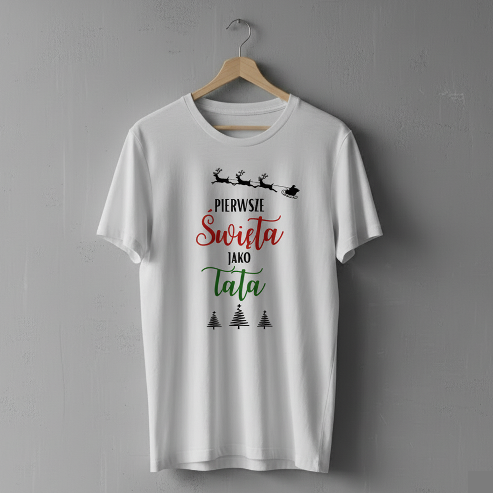 Biała koszulka wisząca na drewnianym wieszaku, ozdobiona nadrukiem z reniferami ciągnącymi sanie oraz napisem ‘Pierwsze Święta jako Tata’ i trzema choinkami.
