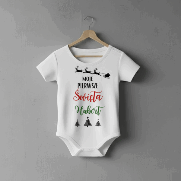 Białe body niemowlęce wiszące na wieszaku, ozdobione nadrukiem z reniferami ciągnącymi sanie oraz napisem ‘Moje pierwsze Święta – Hubert’ i trzema choinkami